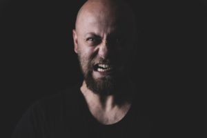 Man with beard and shaved head snarling at the camera in a menacing fashion. A visual representation of Sexual Aggressiveness.