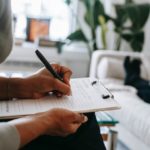 Photo of a person writing on a notepad attached to a clipboard next to a person laying on a couch. Therapy.